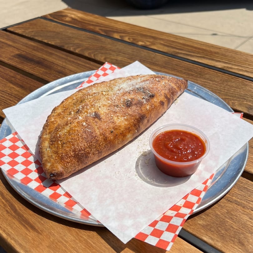 Golden-brown calzone sprinkled with parmesan cheese, served with a side of marinara sauce on a wooden table.